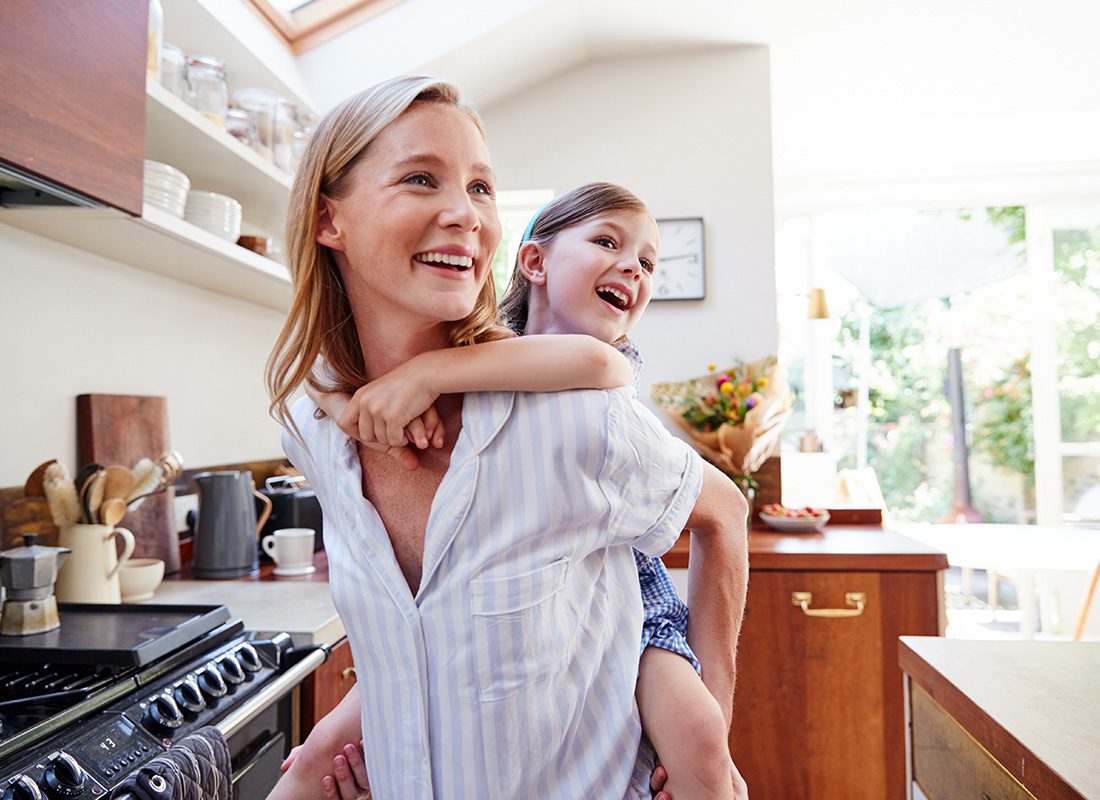 Contact - Portrait of a Smiling Young Mother Giving her Excited Daughter a Piggyback Ride While They Stand in the Kitchen