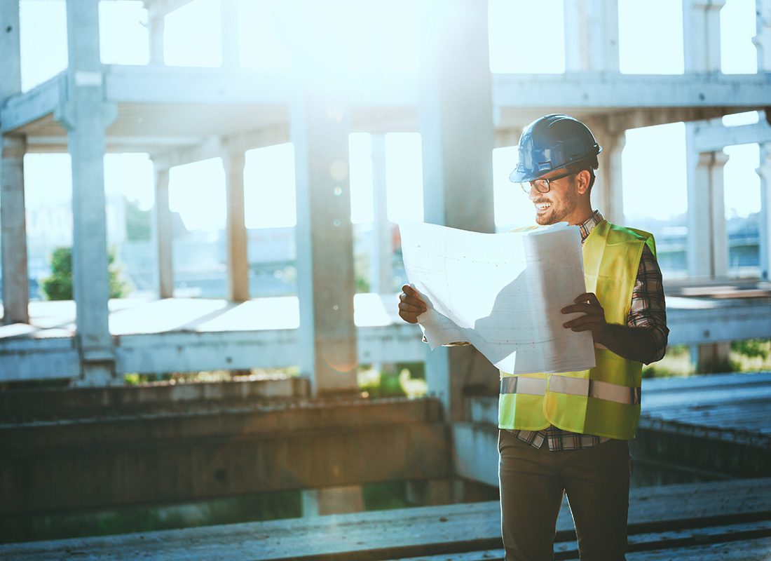 Insurance by Industry - Smiling Young Construction Worker Standing in a Framed Commercial Building Construction Jobsite While Holding and Looking at Blueprints in his Hands