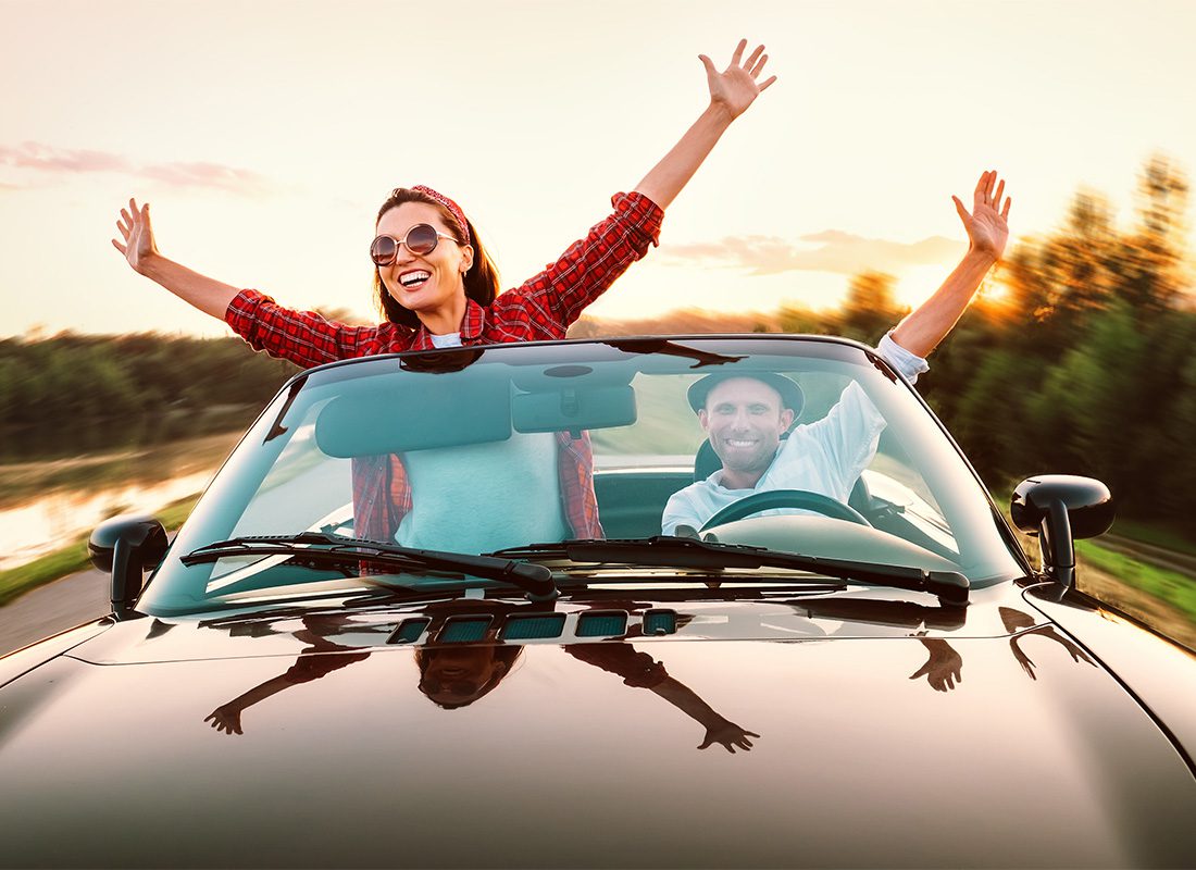 Personal Insurance - View of an Excited Young Couple Sticking Their Hands Out as They Have Fun Driving in a Convertible at Sunset During a Road Trip